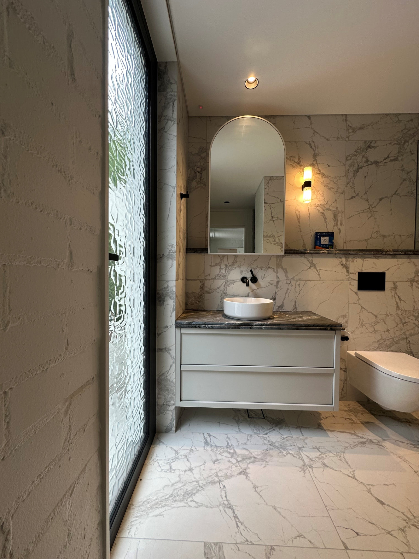 Marble-tiled powder room with floating vanity, dark stone top, vessel basin and arched mirror in Cremorne by Mr Woodworx