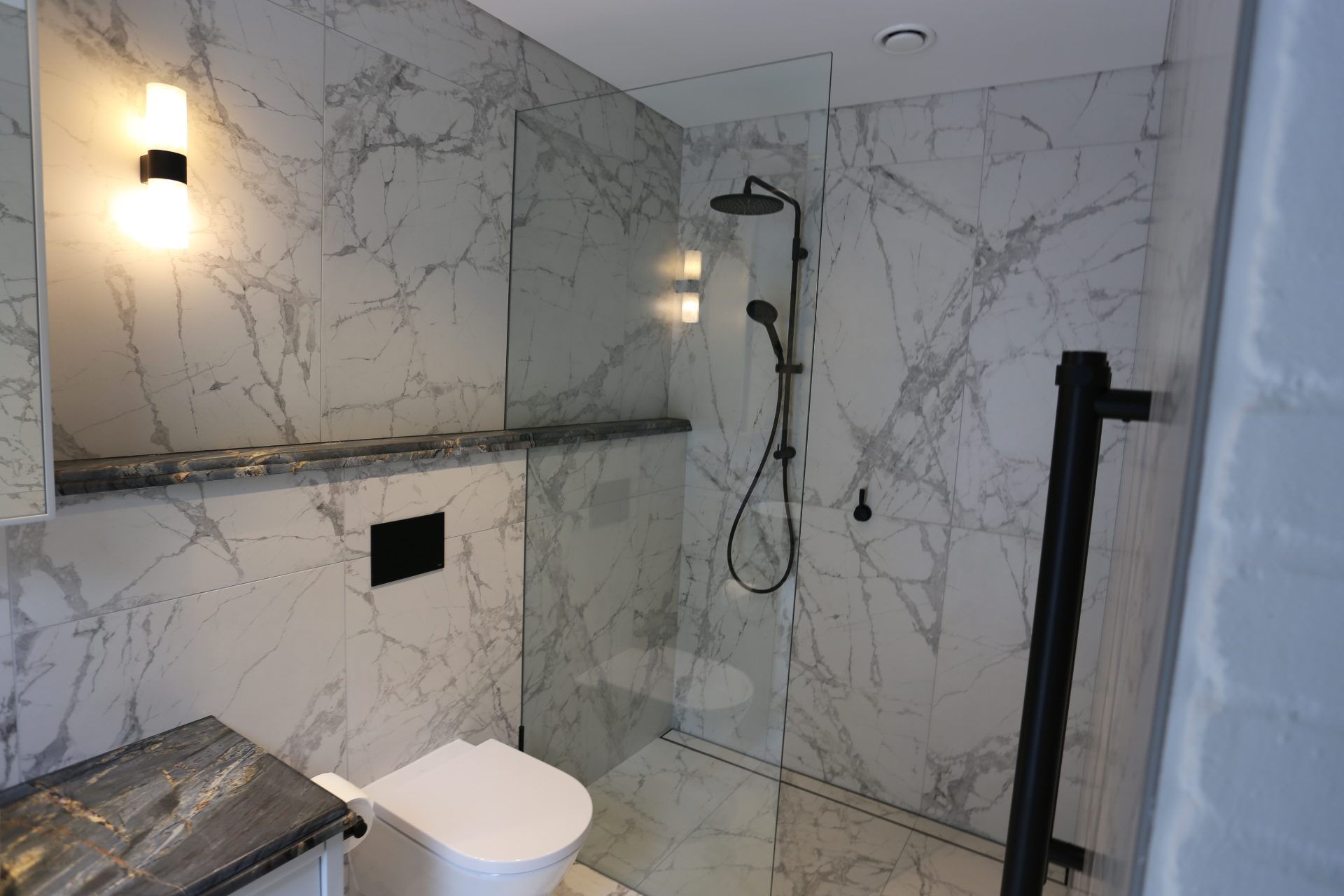 Bathroom with marble-look tiling, frameless shower screen and black fixtures by Mr Woodworx